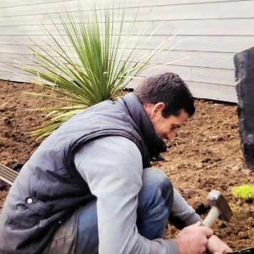 Un homme travaille dans un jardin, utilisant un marteau près de plantations.